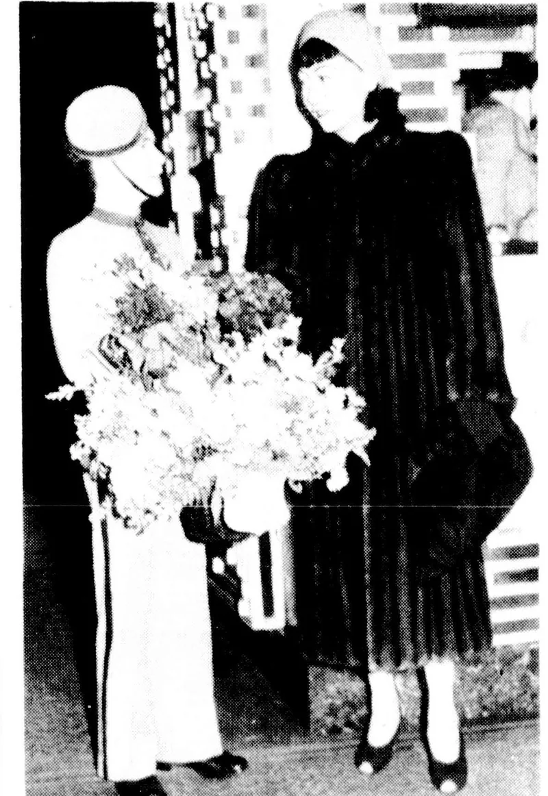 Anna May Wong in a long dark fur coat and close turban taking a large bouquet from a uniformed page boy.