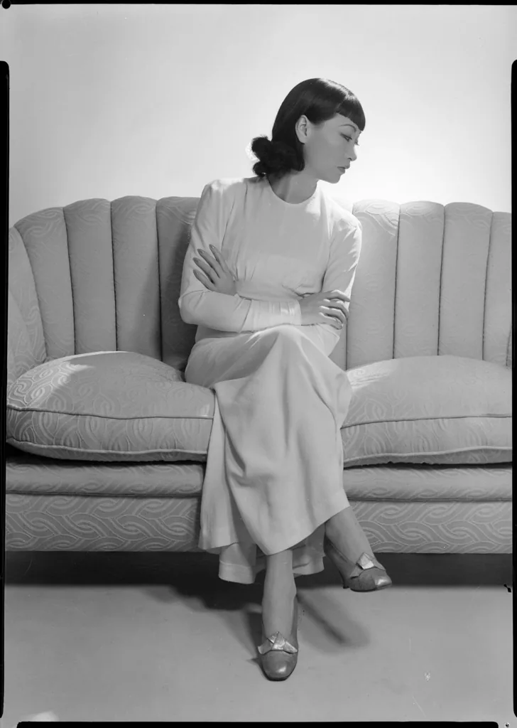 Anna May Wong seated on a channel-backed sofa in a long pale dress, arms folded, looking down to one side.