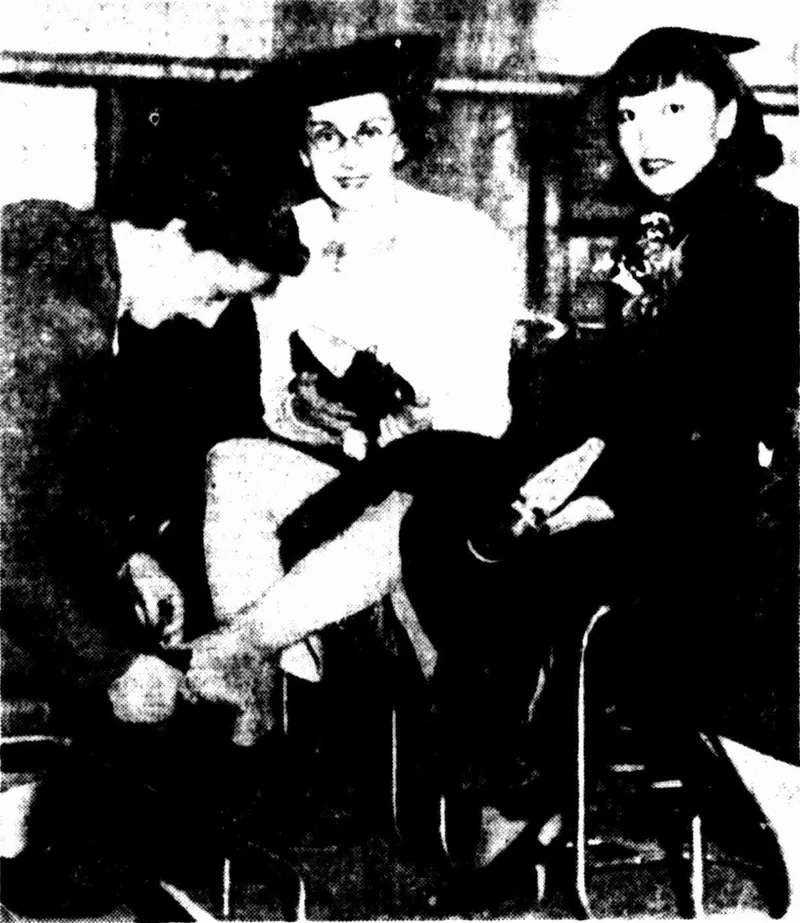 In a shoe department: Anna May Wong seated at right with a pale shoe in her lap, beside a woman in spectacles being fitted by a kneeling assistant.