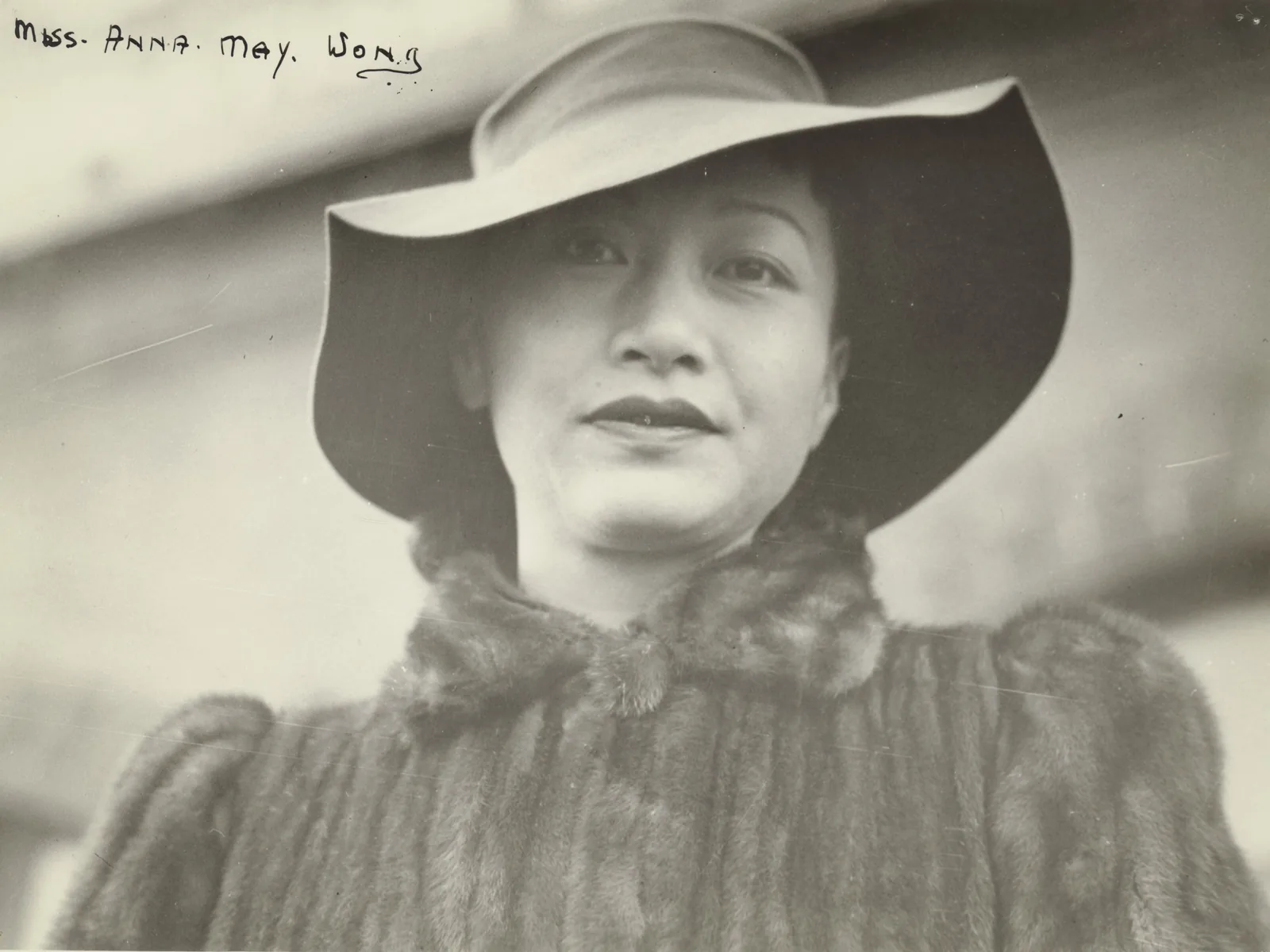 Anna May Wong in a wide-brimmed felt hat and a mink coat, looking down into the camera with a half-smile. Written in ink across the top corner of the print: Miss Anna May Wong.
