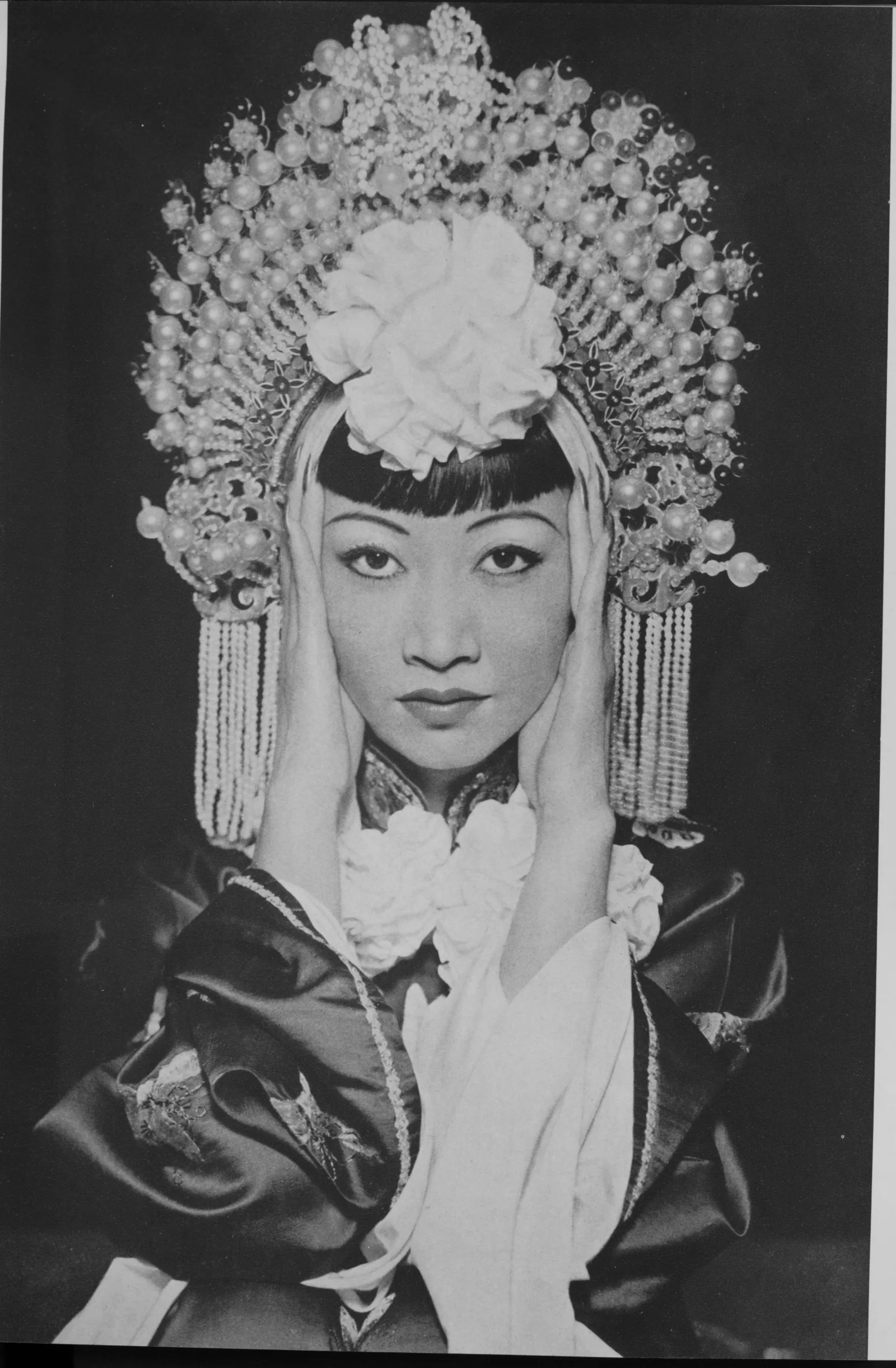 Anna May Wong in a tall pearl headdress with a white silk flower and bead tassels, her palms pressed to her cheeks, looking straight at the camera.