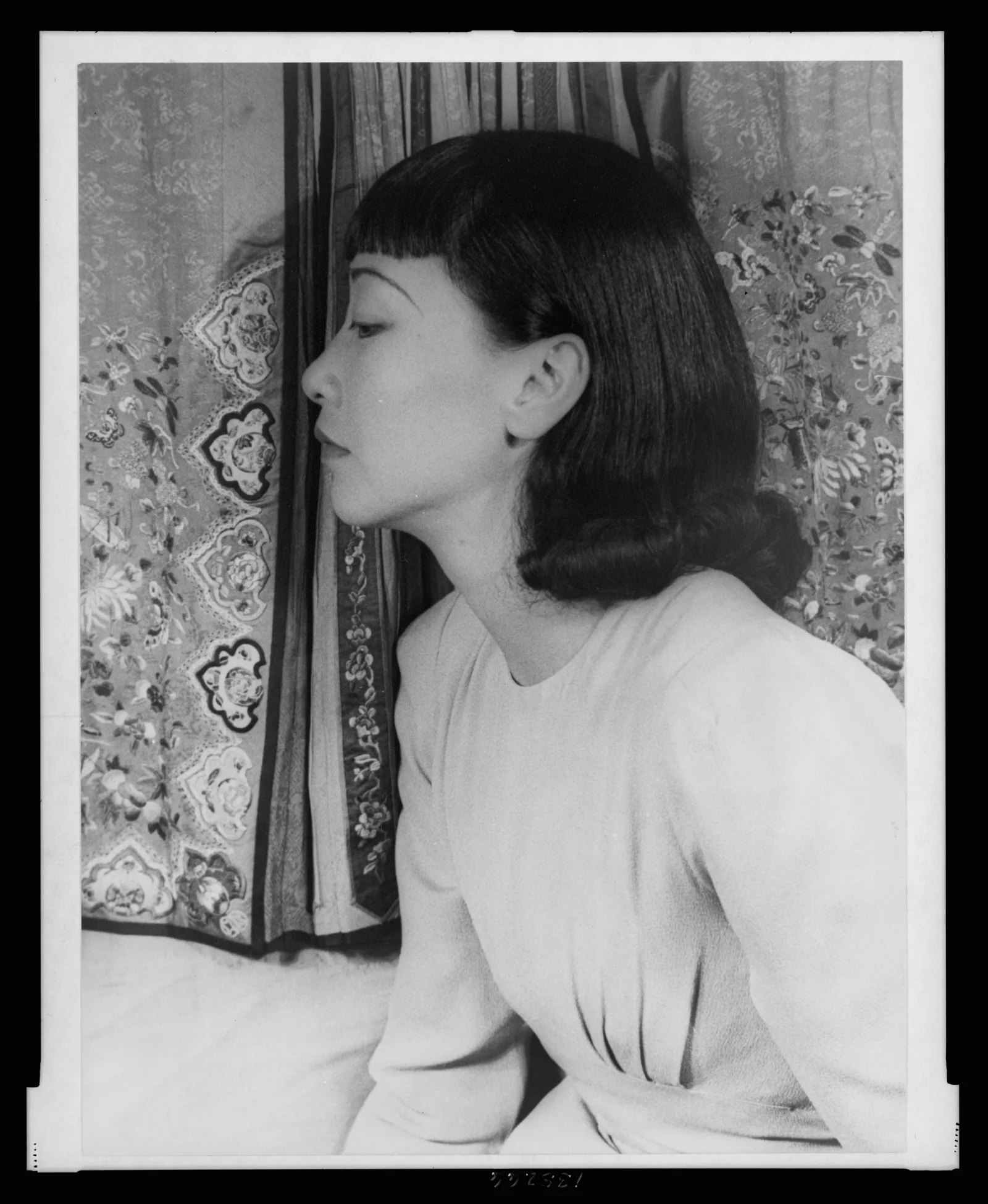 Anna May Wong in profile, eyes lowered, in a pale crepe dress, before embroidered silk panels.
